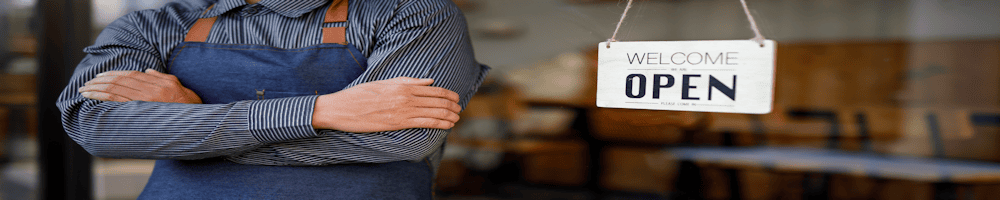 smallbusiness-1000-3 - Newcastle SEO Person in a blue apron and striped shirt stands with arms crossed in front of a shop window, an 'OPEN' sign visible beside them.